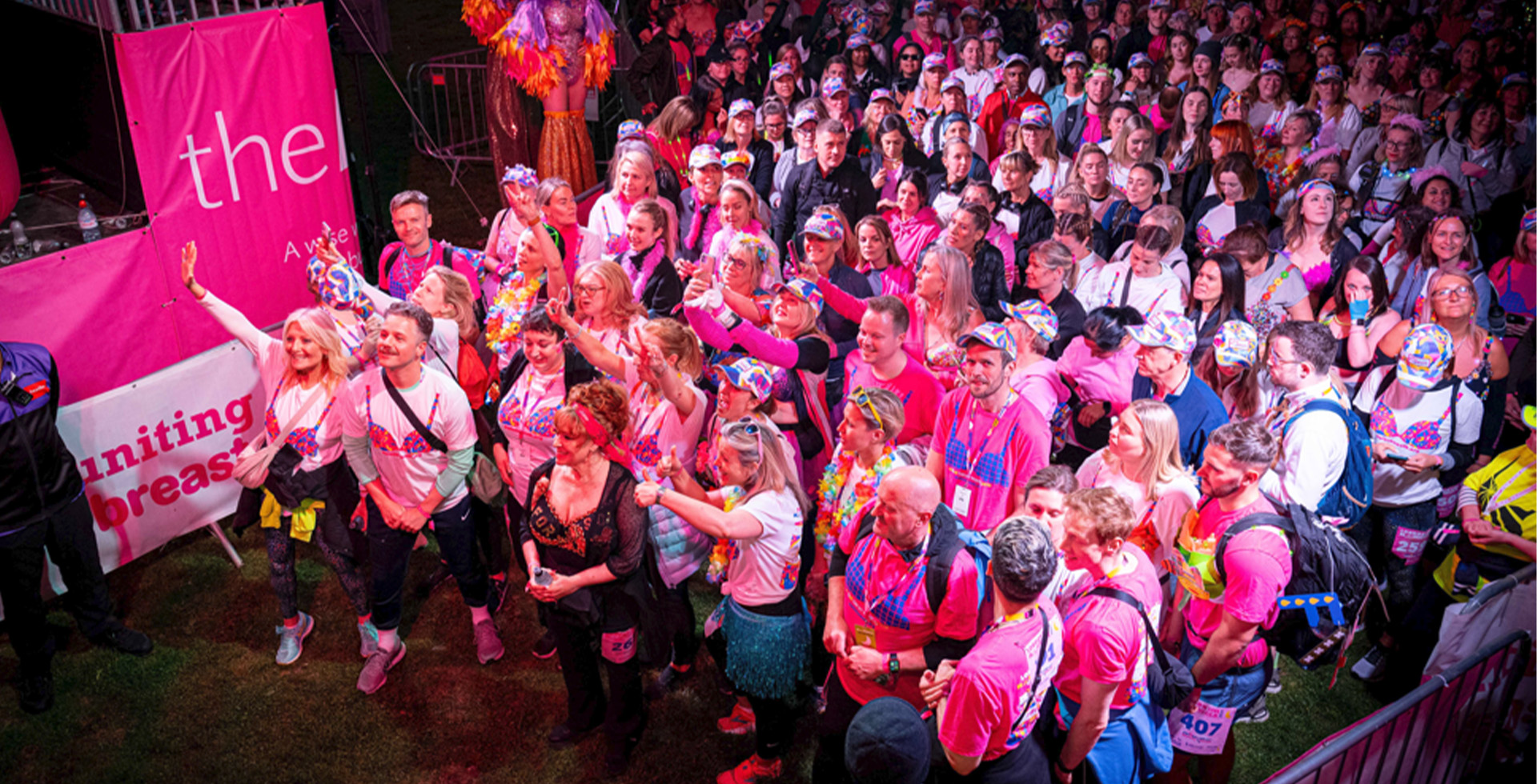 Large crowd of participants gathered at a vibrant nighttime event, dressed in pink and colourful outfits, symbolising unity for a cause. The scene is illuminated by bright lighting, with performers on stilts in the background and signage promoting a breast cancer awareness campaign.