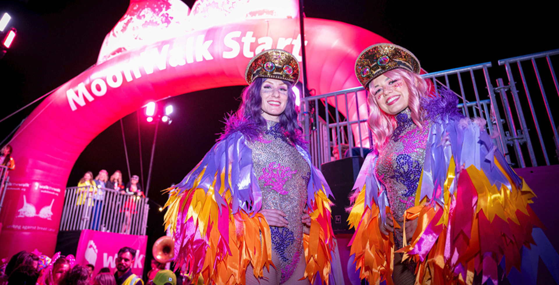 Two vibrant performers dressed in glittering, colorful costumes with feathered accents stand smiling under a glowing pink 'MoonWalk Start' arch. The nighttime event is illuminated by bright lights, with spectators visible in the background, creating an atmosphere of celebration and energy.