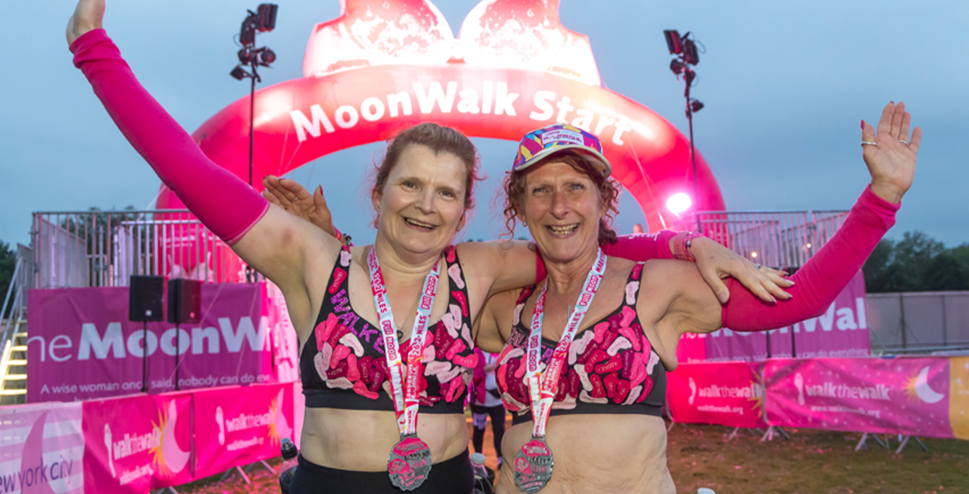 Two joyful participants wearing vibrant bras adorned with pink designs and medals around their necks pose proudly under the illuminated 'MoonWalk Start' arch. The bright and colorful backdrop highlights the celebratory spirit of the event.